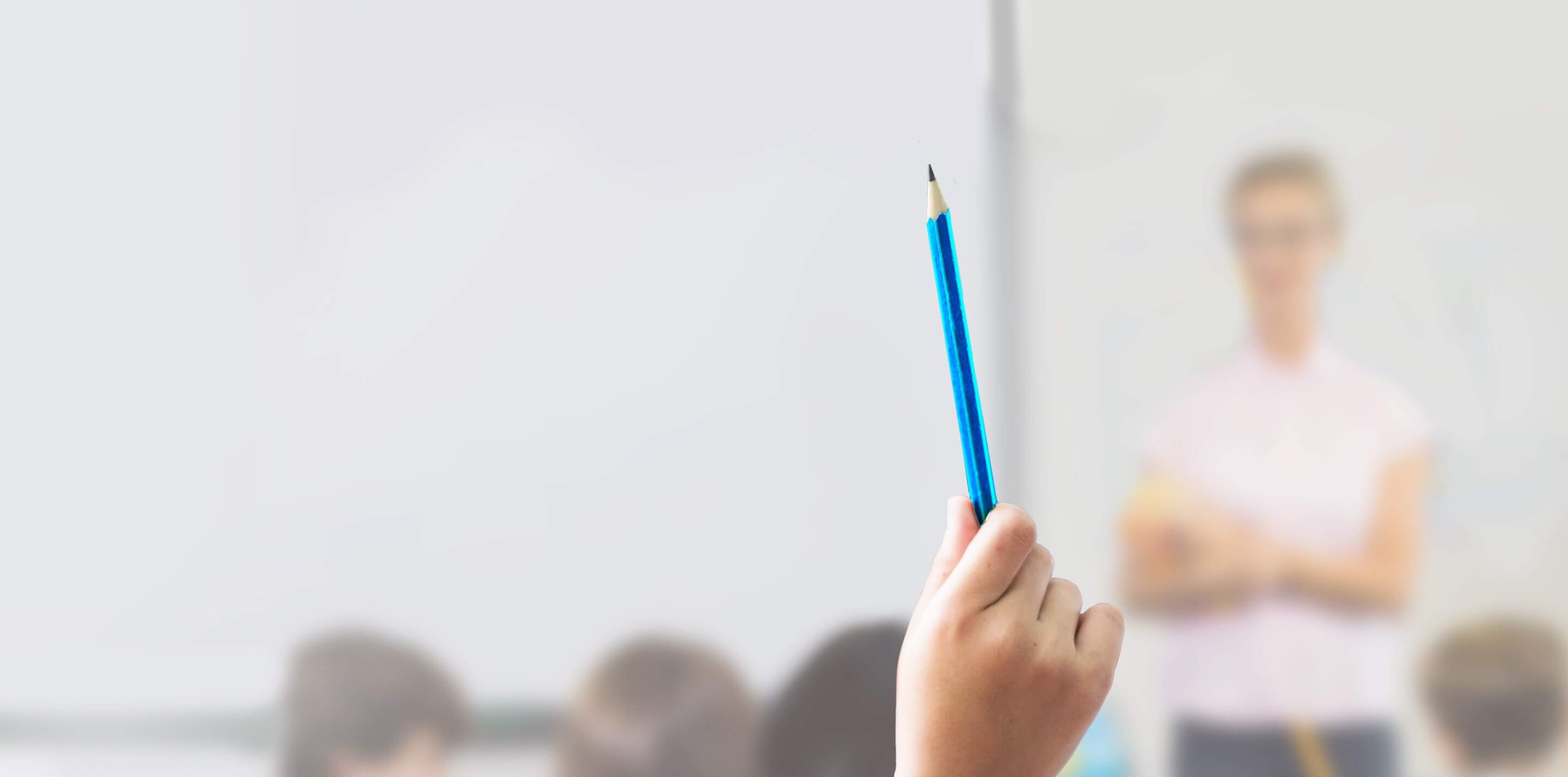 Hand raised holding pencil in a classroom with a teacher